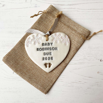 Personalised bumps first Christmas heart ornament, pearlised white clay with BABY ROBINSON DUE 2026 painted grey, decorated with 2 iridescent glitter snowflakes on either side of the top of the heart and baby feet cut out at the bottom