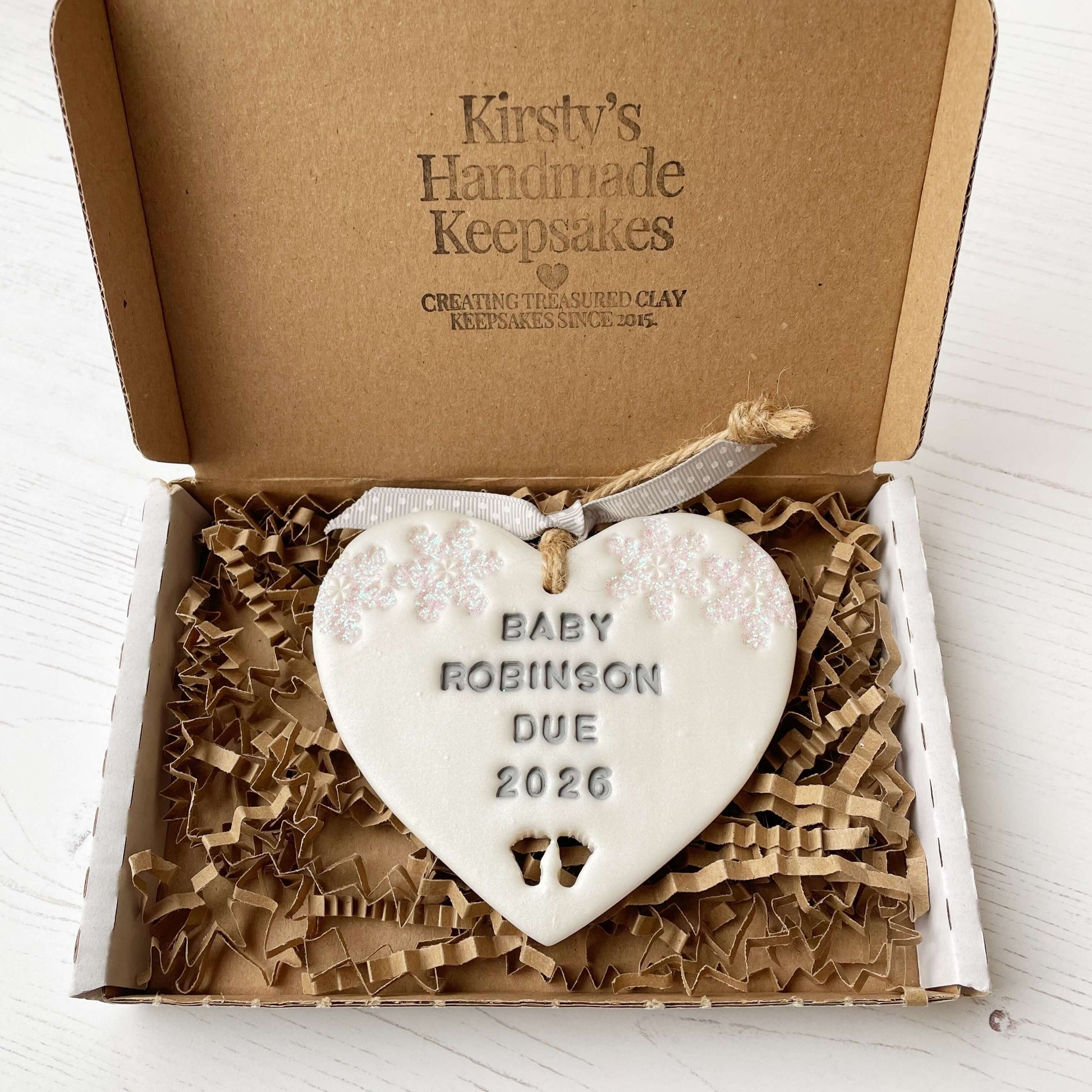 Personalised bumps first Christmas heart ornament, pearlised white clay with BABY ROBINSON DUE 2026 painted grey, decorated with 2 iridescent glitter snowflakes on either side of the top of the heart and baby feet cut out at the bottom