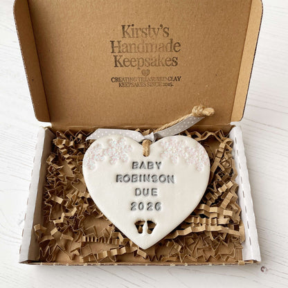 Personalised bumps first Christmas heart ornament, pearlised white clay with BABY ROBINSON DUE 2026 painted grey, decorated with 2 iridescent glitter snowflakes on either side of the top of the heart and baby feet cut out at the bottom
