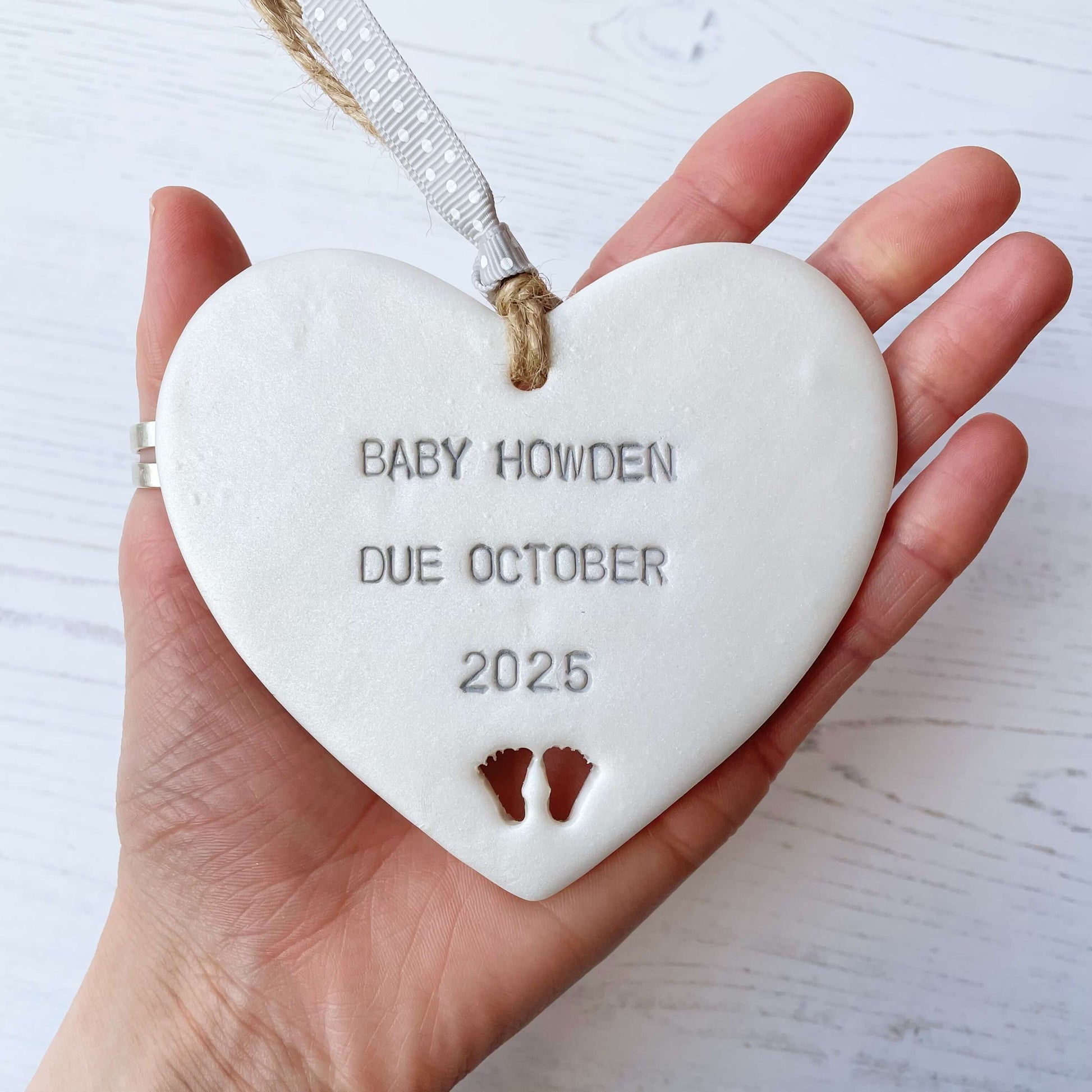 Pearlised white clay hanging heart with baby feet cut out of the bottom and grey personalisation, the heart is personalised with BABY HOWDEN DUE OCTOBER 2025