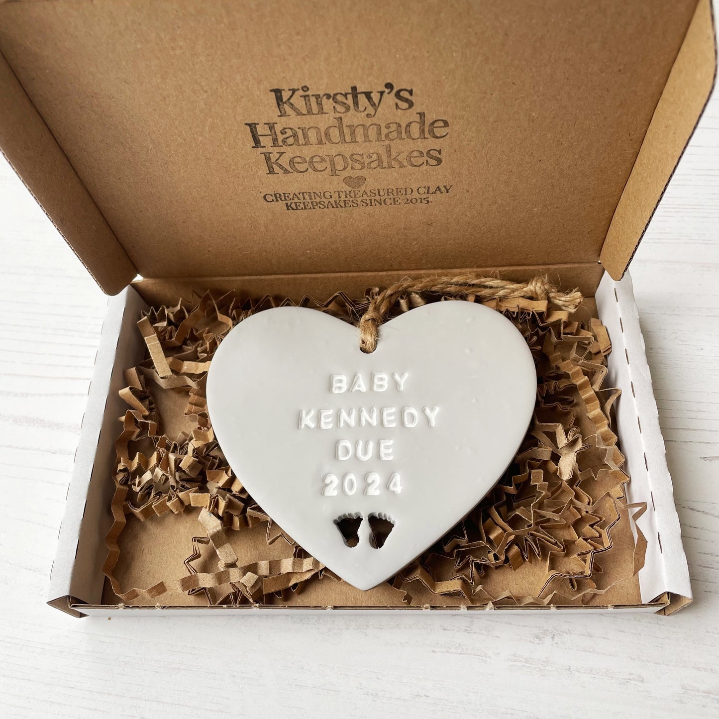 Personalised pregnancy reveal sign keepsake, grey clay hanging heart with baby feet cut out at the bottom, the heart is personalised in white with BABY KENNEDY DUE 2024 In a white postal box with brown Kraft shredded zigzag paper
