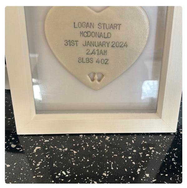 A personalised pearlised white polymer clay heart keepsake plaque inside a white wooden frame, with custom baby birth details printed on the front, displayed on a black speckled countertop.