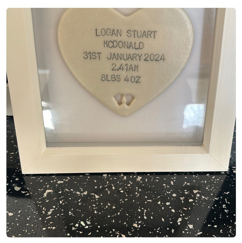 A personalised pearlised white polymer clay heart keepsake plaque inside a white wooden frame, with custom baby birth details printed on the front, displayed on a black speckled countertop.