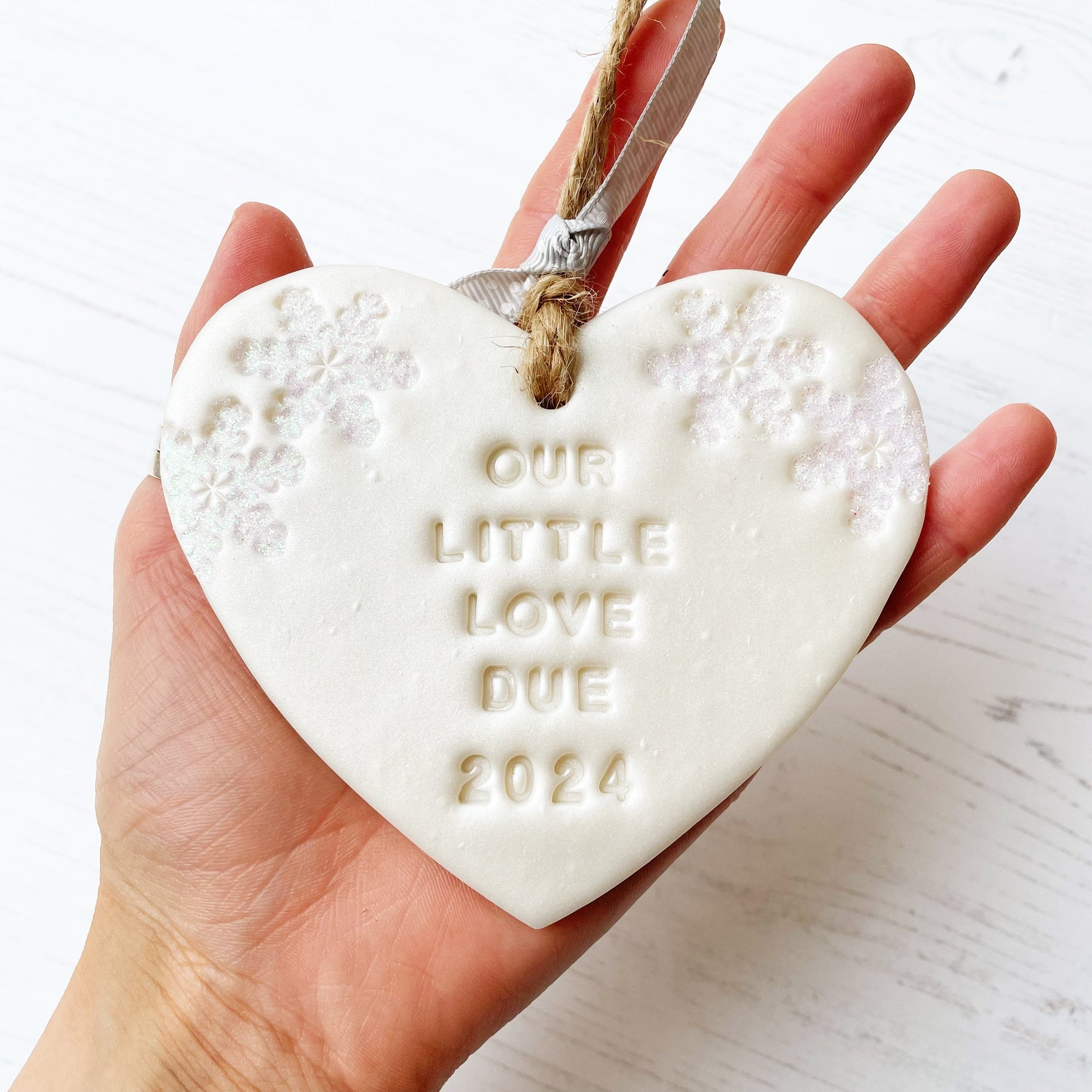 Personalised baby reveal Christmas heart ornament, pearlised white clay with OUR LITTLE LOVE DUE 2024 (text not painted), decorated with 2 iridescent glitter snowflakes on either side of the top of the heart