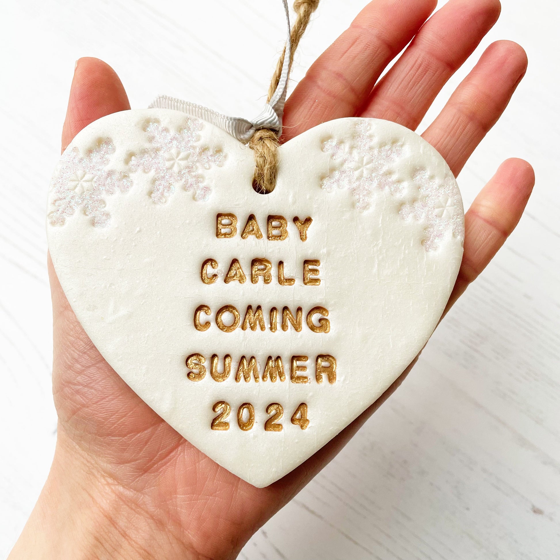 Personalised baby's first Christmas heart ornament, pearlised white clay with BABY CARLE DUE SUMMER 2024 painted gold, decorated with 2 iridescent glitter snowflakes on either side of the top of the heart