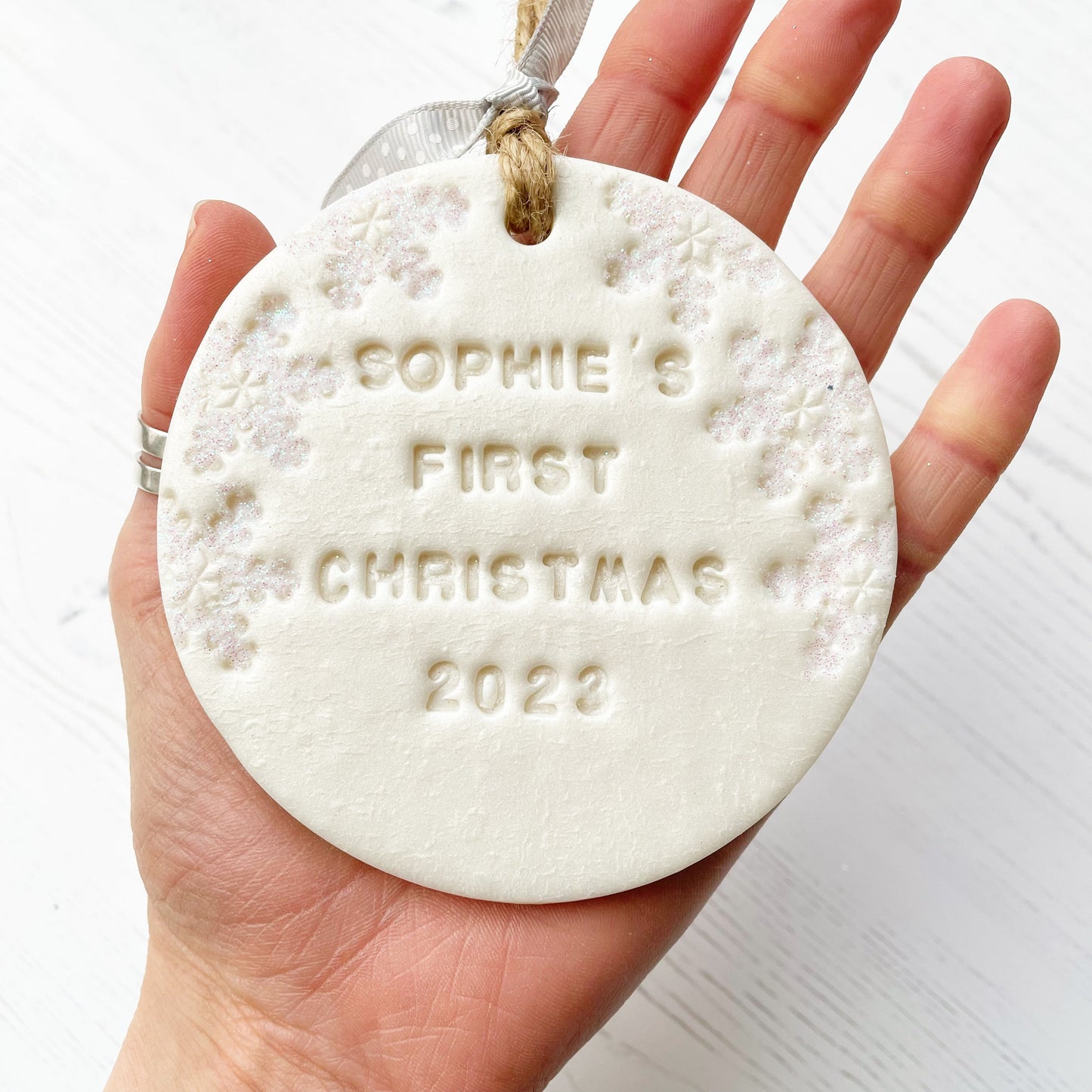 Personalised baby’s first Christmas bauble ornament, pearlised white round clay with SOPHIE’S FIRST CHRISTMAS 2023 (letters not painted), decorated with 3 iridescent glitter snowflakes on either side of the bauble