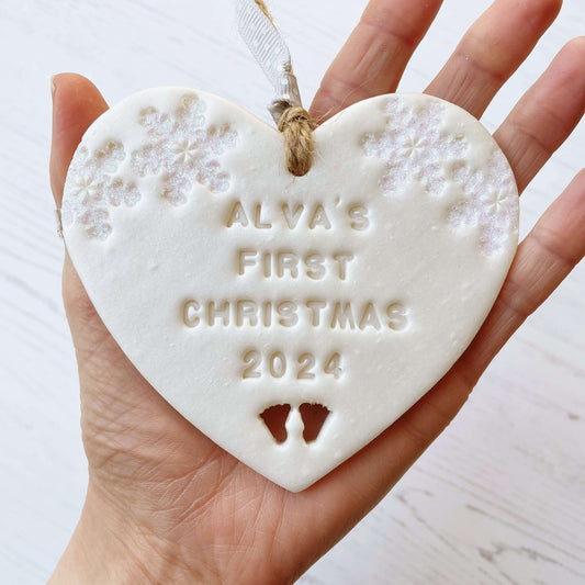 Personalised baby’s first Christmas heart ornament, pearlised white clay with ALVA’S FIRST CHRISTMAS 2024, decorated with 2 iridescent glitter snowflakes on either side of the top of the heart and baby feet cut out at the bottom