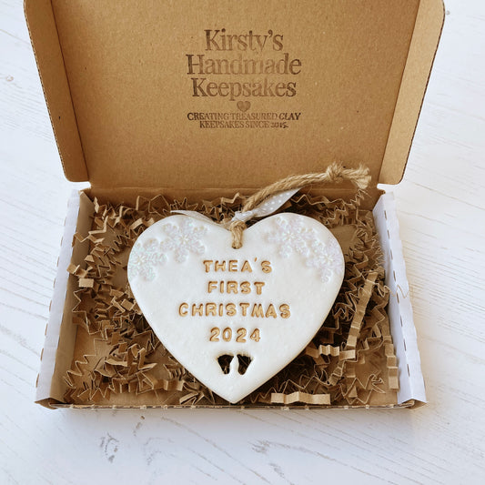 Personalised baby’s first Christmas heart ornament, pearlised white clay with THEA’S FIRST CHRISTMAS 2024 painted gold, decorated with 2 iridescent glitter snowflakes on either side of the top of the heart and baby feet cut out at the bottom