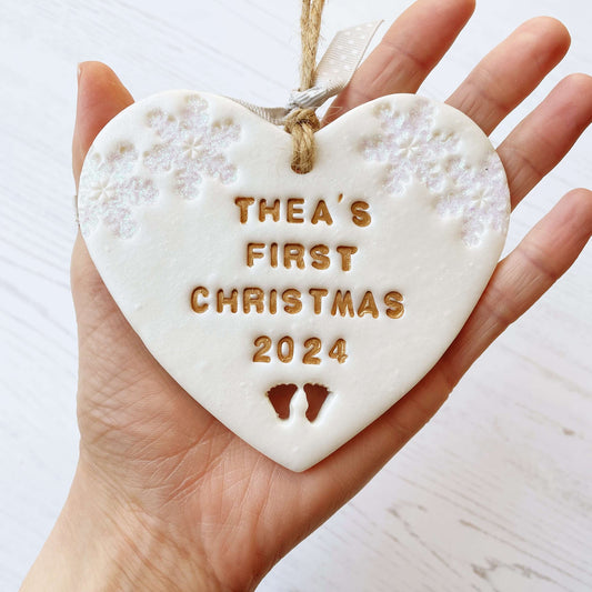 Personalised baby’s first Christmas heart ornament, pearlised white clay with THEA’S FIRST CHRISTMAS 2024 painted gold, decorated with 2 iridescent glitter snowflakes on either side of the top of the heart and baby feet cut out at the bottom