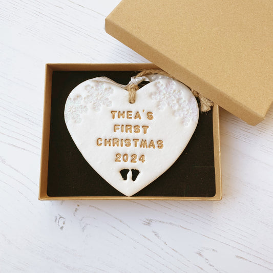 Personalised baby’s first Christmas heart ornament, pearlised white clay with THEA’S FIRST CHRISTMAS 2024 painted gold, decorated with 2 iridescent glitter snowflakes on either side of the top of the heart and baby feet cut out at the bottom