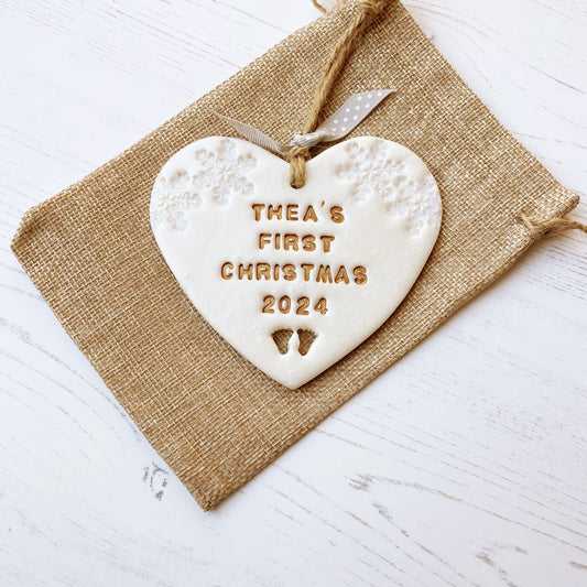 Personalised baby’s first Christmas heart ornament, pearlised white clay with THEA’S FIRST CHRISTMAS 2024 painted gold, decorated with 2 iridescent glitter snowflakes on either side of the top of the heart and baby feet cut out at the bottom