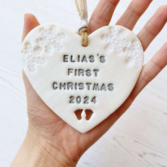 Personalised baby’s first Christmas heart ornament, pearlised white clay with ELIAS’S FIRST CHRISTMAS 2024 painted grey, decorated with 2 iridescent glitter snowflakes on either side of the top of the heart and baby feet cut out at the bottom