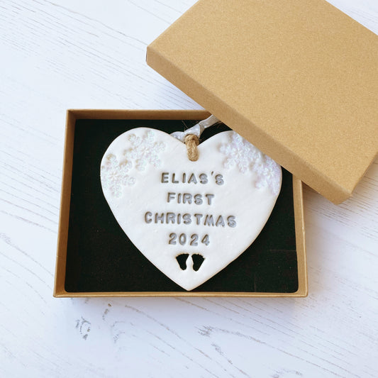 Personalised baby’s first Christmas heart ornament, pearlised white clay with ELIAS’S FIRST CHRISTMAS 2024 painted grey, decorated with 2 iridescent glitter snowflakes on either side of the top of the heart and baby feet cut out at the bottom