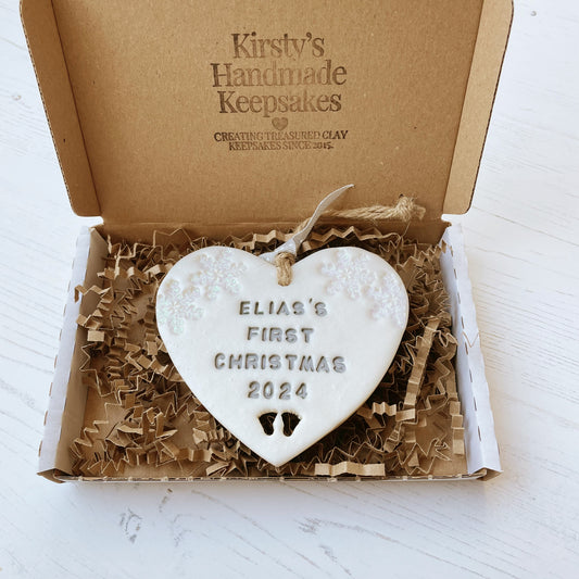 Personalised baby’s first Christmas heart ornament, pearlised white clay with ELIAS’S FIRST CHRISTMAS 2024 painted grey, decorated with 2 iridescent glitter snowflakes on either side of the top of the heart and baby feet cut out at the bottom