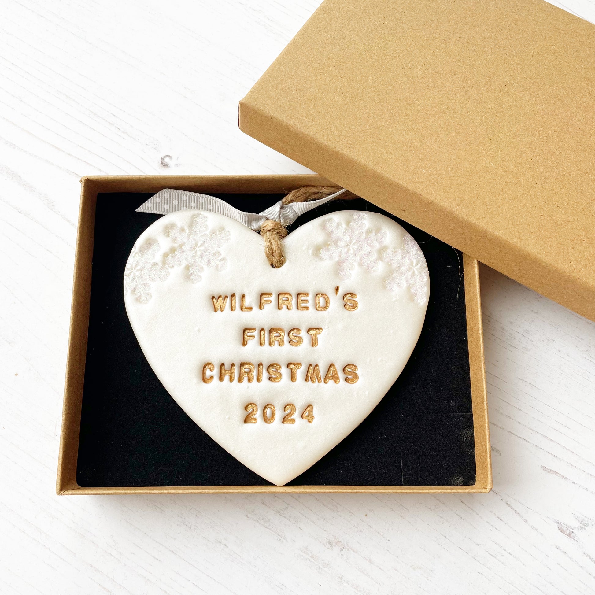 Personalised baby’s first Christmas heart ornament, pearlised white clay with WILFRED’S FIRST CHRISTMAS 2024 painted gold, decorated with 2 iridescent glitter snowflakes on either side of the top of the heart In a brown Kraft luxury gift box