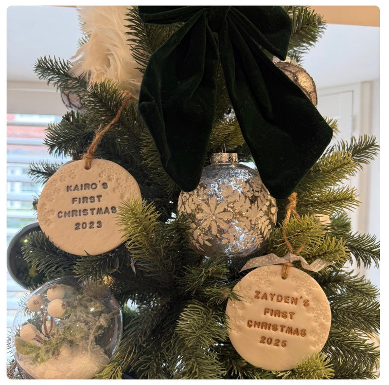Personalised round pearlised white polymer clay Christmas ornaments hanging on a decorated tree, with one ornament reading 'KAIRO'S FIRST CHRISTMAS 2023' and another reading 'ZAYDEN'S FIRST CHRISTMAS 2025'.