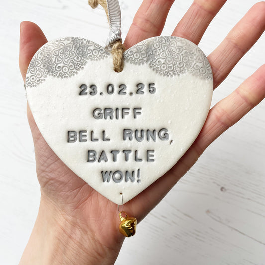 Personalised cancer survivor gift, pearlised white clay hanging heart with a grey lace edge at the top of the heart and a gold bell hanging below, the heart is personalised with 23.02.25 GRIFF BELL RAUNG BATTLE WON