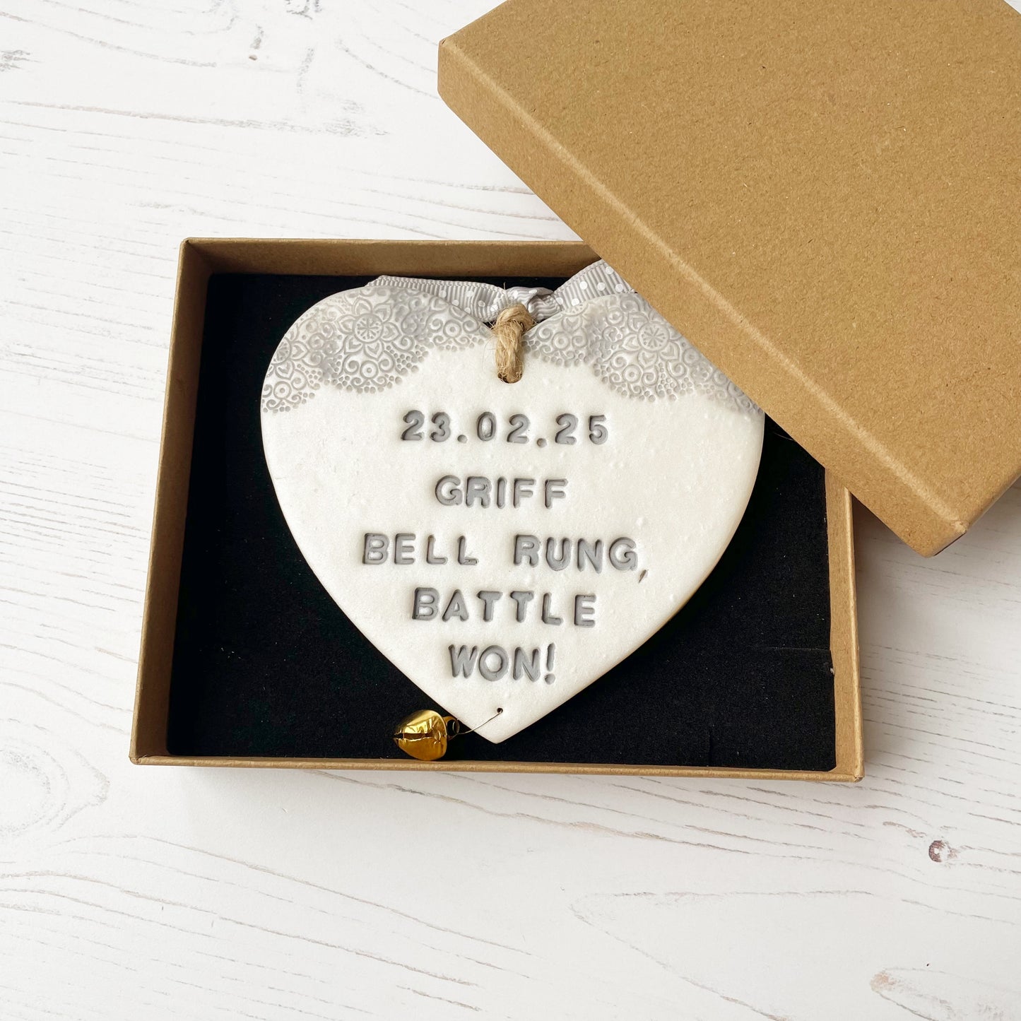 Personalised cancer survivor gift, pearlised white clay hanging heart with a grey lace edge at the top of the heart and a gold bell hanging below, the heart is personalised with 23.02.25 GRIFF BELL RAUNG BATTLE WON