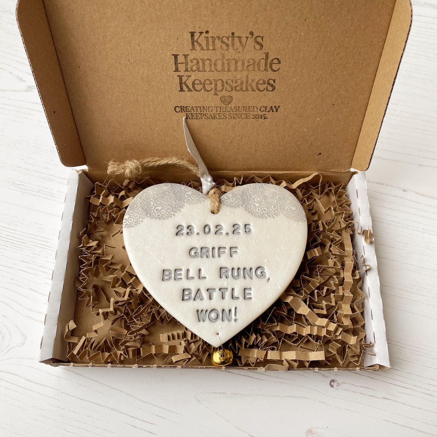 Personalised cancer survivor gift, pearlised white clay hanging heart with a grey lace edge at the top of the heart and a gold bell hanging below, the heart is personalised with 23.02.25 GRIFF BELL RAUNG BATTLE WON