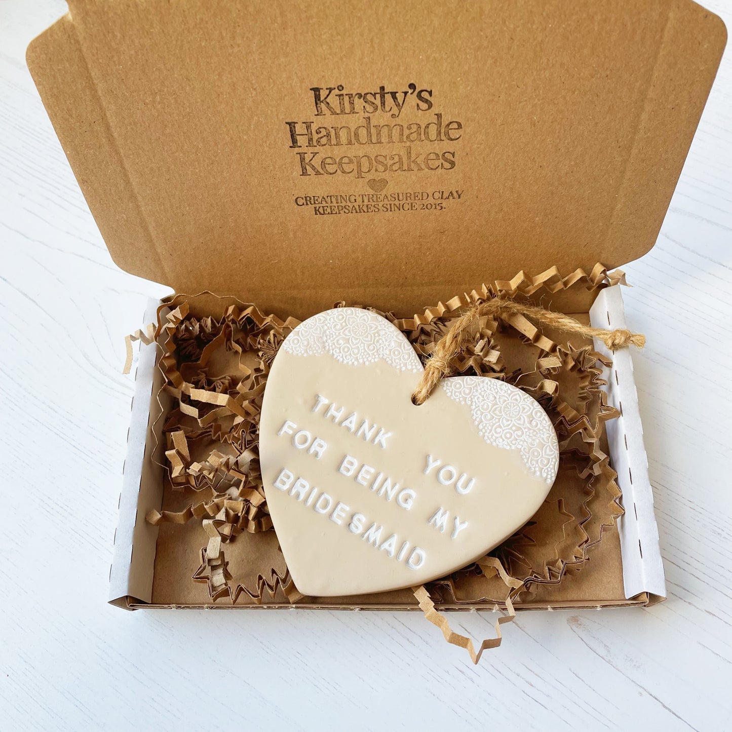 Personalised Bridesmaid thank you gift, beige clay hanging heart with a white lace edge at the top of the heart, the heart is personalised with THANK YOU FOR BEING MY BRIDESMAID In a postal box with Kraft brown shredded zigzag paper