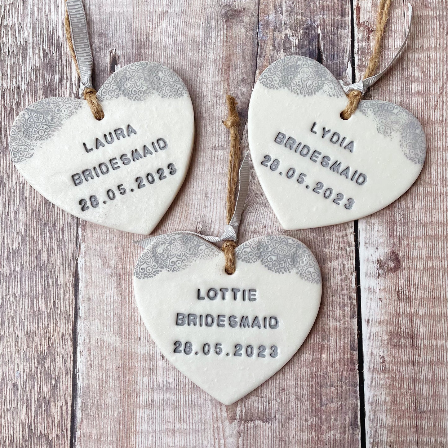 3 Personalised Bridesmaid thank you gifts, pearlised white clay hanging hearts with a grey lace edge at the top of the heart, the hearts are personalised with LAURA BRIDESMAID 28.05.2023, LYDIA BRIDESMAID 28.05.2023 and LOTTIE BRIDESMAID 28.05.2023
