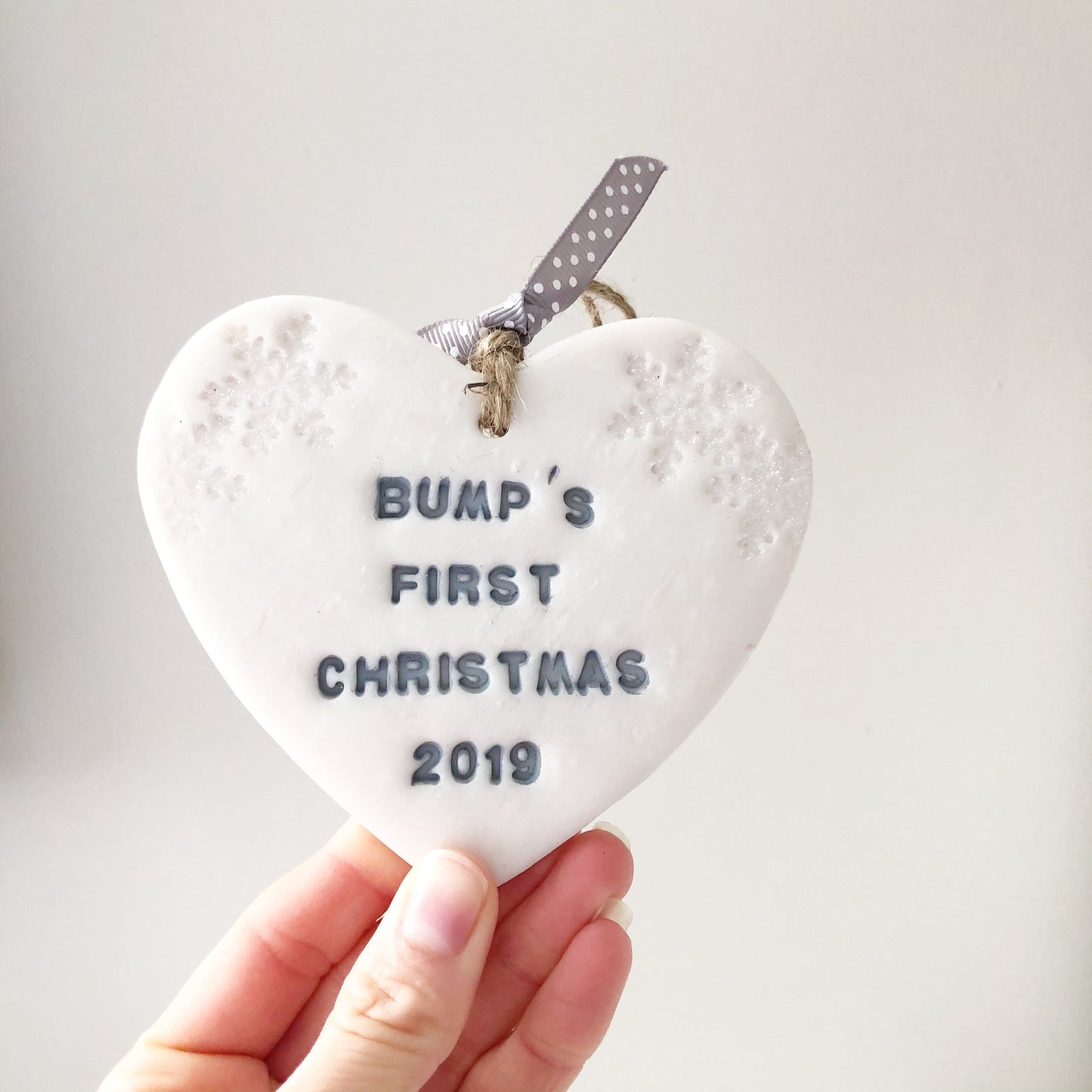 Personalised bump's first Christmas heart ornament, pearlised white clay with BUMP'S FIRST CHRISTMAS 2019 painted grey, decorated with 2 iridescent glitter snowflakes on either side of the top of the heart