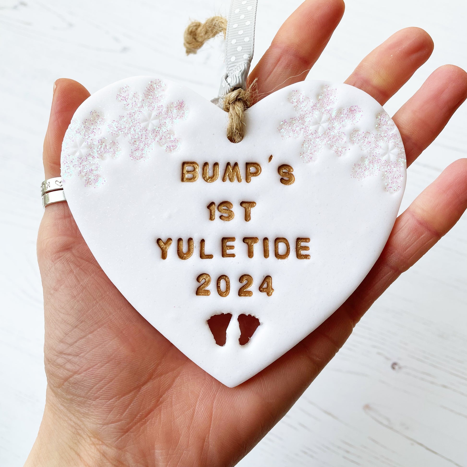 Personalised bump’s first Christmas heart ornament, glitter white clay with BUMP’S 1ST YULETIDE 2024 painted gold, decorated with 2 iridescent glitter snowflakes on either side of the top of the heart and baby feet cut out at the bottom