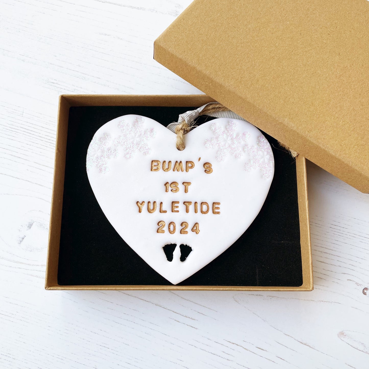 Personalised bump’s first Christmas heart ornament, glitter white clay with BUMP’S 1ST YULETIDE 2024 painted gold, decorated with 2 iridescent glitter snowflakes on either side of the top of the heart and baby feet cut out at the bottom