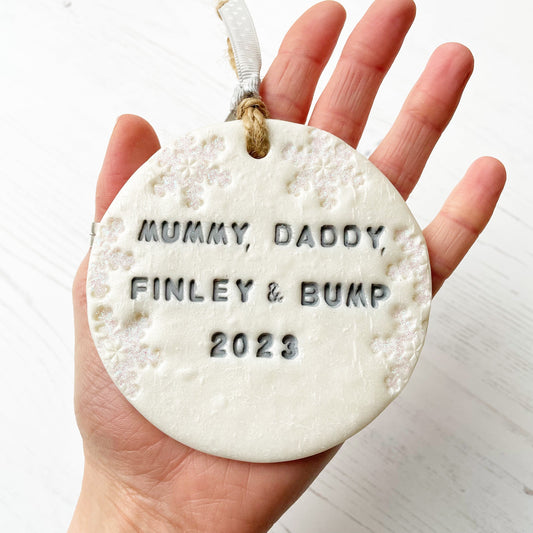 Personalised bump’s first Christmas bauble ornament, pearlised white round clay with MUMMY, DADDY, FINLEY & BUMP CHRISTMAS 2023 in grey paint, decorated with 3 iridescent glitter snowflakes on either side of the bauble