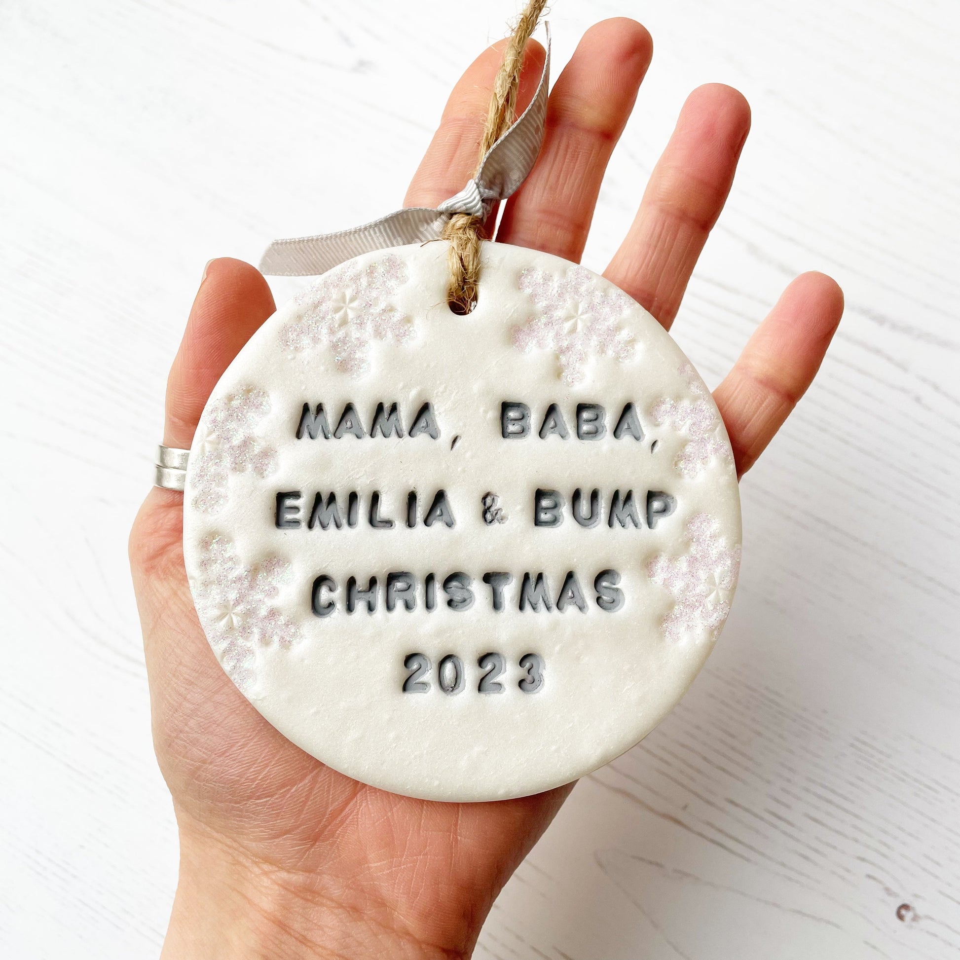 Personalised bump’s first Christmas bauble ornament, pearlised white round clay with MAMA, BABA, EMILIA & BUMP CHRISTMAS 2023 in grey paint, decorated with 3 iridescent glitter snowflakes on either side of the bauble