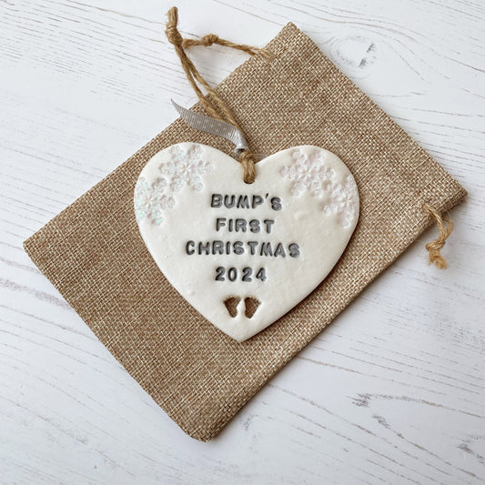 Personalised bump’s first Christmas heart ornament, pearlised white clay with BUMP’S FIRST CHRISTMAS 2024 painted grey, decorated with 2 iridescent glitter snowflakes on either side of the top of the heart and baby feet cut out at the bottom