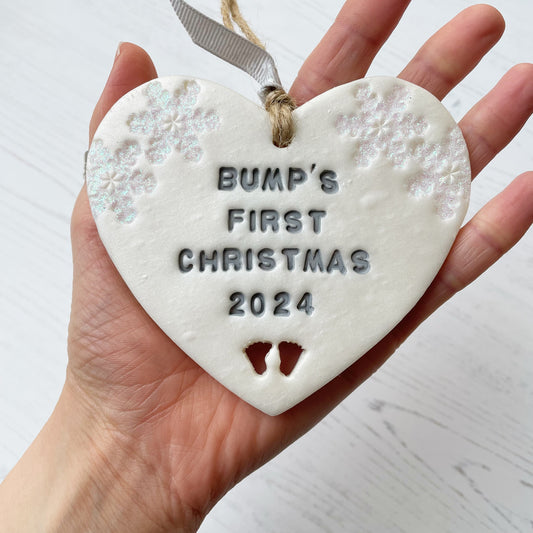 Personalised bump’s first Christmas heart ornament, pearlised white clay with BUMP’S FIRST CHRISTMAS 2024 painted grey, decorated with 2 iridescent glitter snowflakes on either side of the top of the heart and baby feet cut out at the bottom