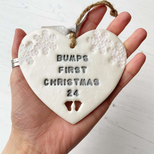 Personalised bump’s first Christmas heart ornament, pearlised white clay with BUMPS FIRST CHRISTMAS 24 painted grey, decorated with 2 iridescent glitter snowflakes on either side of the top of the heart and baby feet cut out at the bottom