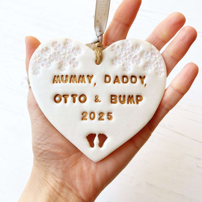 Personalised bumps first Christmas heart ornament, pearlised white clay with MUMMY, DADDY, OTTO & BUMP 2025 painted gold, decorated with 2 iridescent glitter snowflakes on either side of the top of the heart and baby feet cut out at the bottom