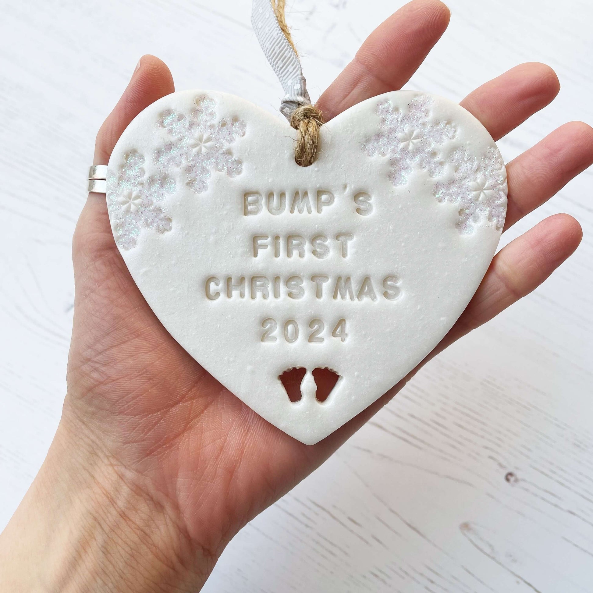 Personalised baby’s first Christmas heart ornament, pearlised white clay with ALVA’S FIRST CHRISTMAS 2024, decorated with 2 iridescent glitter snowflakes on either side of the top of the heart and baby feet cut out at the bottom