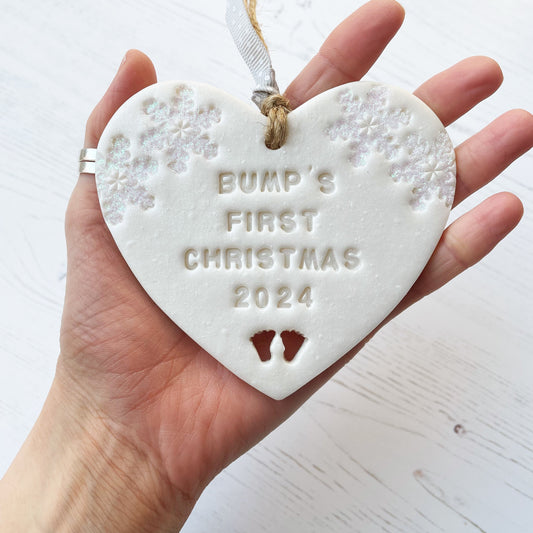 Personalised bump’s first Christmas heart ornament, pearlised white clay with BUMP’S FIRST CHRISTMAS 2024 (not painted), decorated with 2 iridescent glitter snowflakes on either side of the top of the heart and baby feet cut out at the bottom