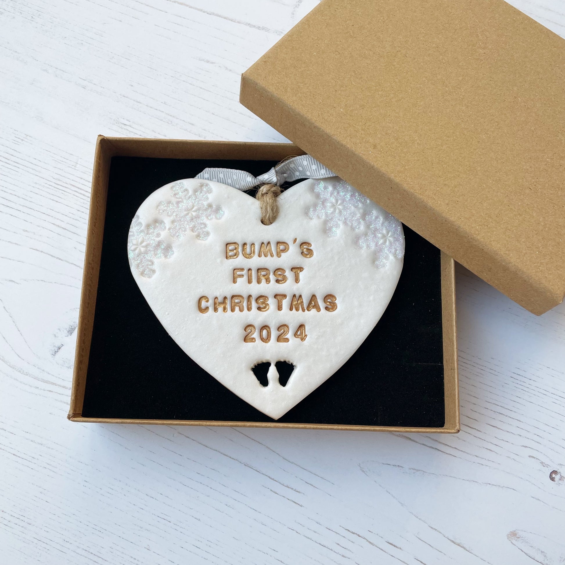 Personalised bump’s first Christmas heart ornament, pearlised white clay with BUMP’S FIRST CHRISTMAS 2024 painted gold, decorated with 2 iridescent glitter snowflakes on either side of the top of the heart and baby feet cut out at the bottom