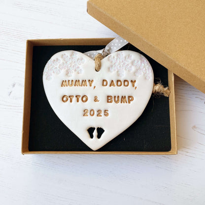 Personalised bumps first Christmas heart ornament, pearlised white clay with MUMMY, DADDY, OTTO & BUMP 2025 painted gold, decorated with 2 iridescent glitter snowflakes on either side of the top of the heart and baby feet cut out at the bottom