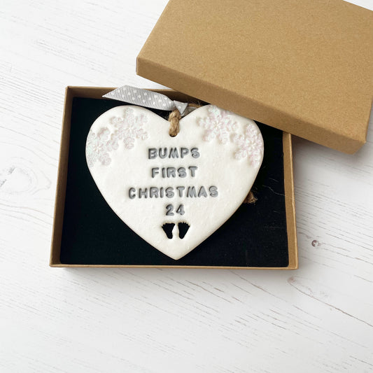 Personalised bump’s first Christmas heart ornament, pearlised white clay with BUMPS FIRST CHRISTMAS 24 painted grey, decorated with 2 iridescent glitter snowflakes on either side of the top of the heart and baby feet cut out at the bottom