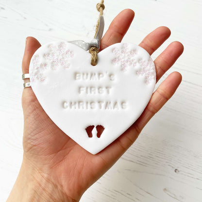 Personalised bump’s first Christmas heart ornament, glitter white clay with BUMP’S FIRST CHRISTMAS, decorated with 2 iridescent glitter snowflakes on either side of the top of the heart and baby feet cut out at the bottom