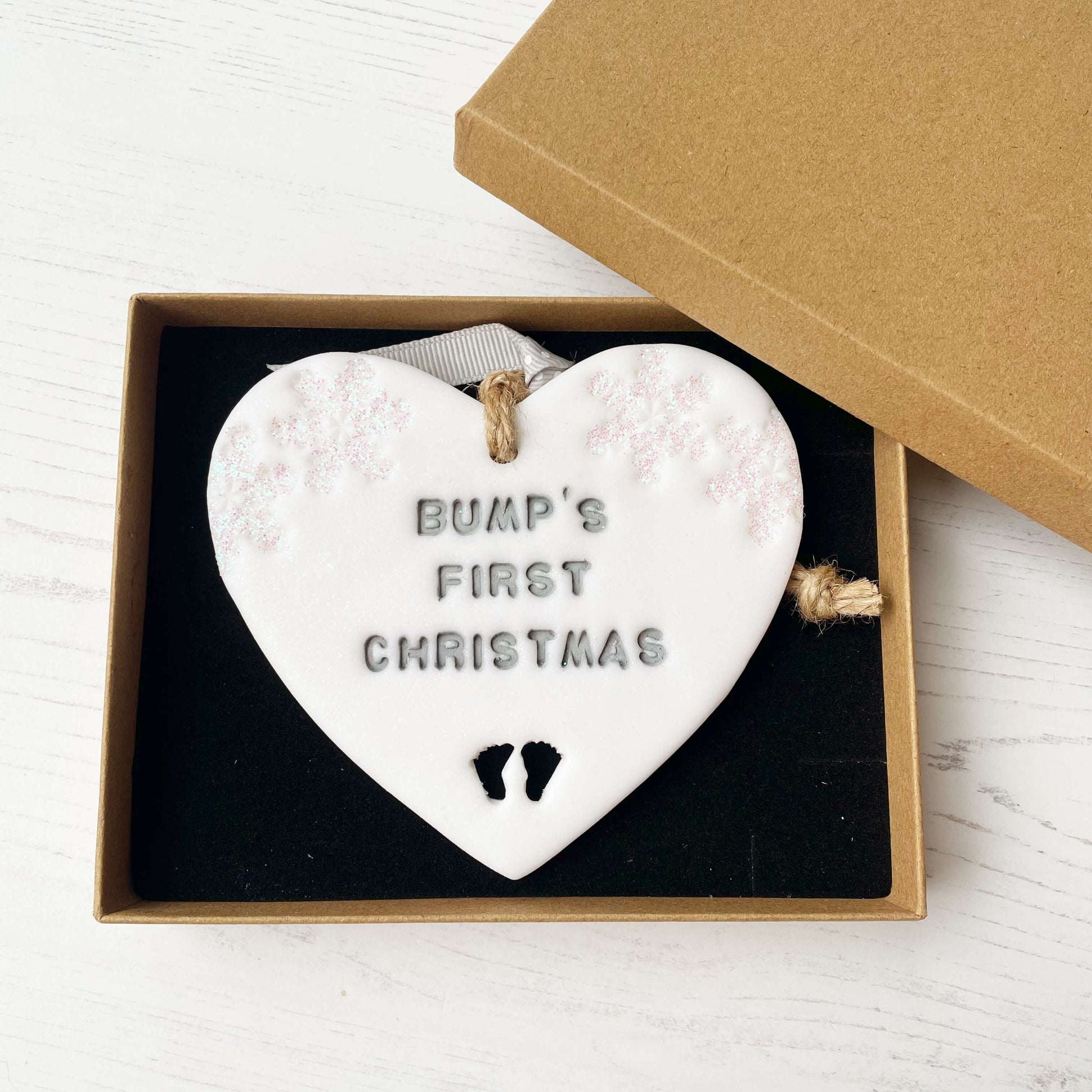 Personalised bump’s first Christmas heart ornament, glitter white clay with BUMP’S FIRST CHRISTMAS painted grey, decorated with 2 iridescent glitter snowflakes on either side of the top of the heart and baby feet cut out at the bottom