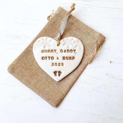 Personalised bumps first Christmas heart ornament, pearlised white clay with MUMMY, DADDY, OTTO & BUMP 2025 painted gold, decorated with 2 iridescent glitter snowflakes on either side of the top of the heart and baby feet cut out at the bottom