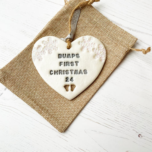 Personalised bump’s first Christmas heart ornament, pearlised white clay with BUMPS FIRST CHRISTMAS 24 painted grey, decorated with 2 iridescent glitter snowflakes on either side of the top of the heart and baby feet cut out at the bottom