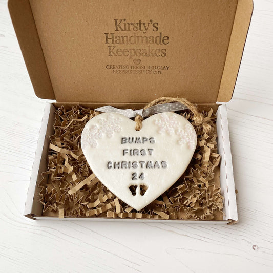 Personalised bump’s first Christmas heart ornament, pearlised white clay with BUMPS FIRST CHRISTMAS 24 painted grey, decorated with 2 iridescent glitter snowflakes on either side of the top of the heart and baby feet cut out at the bottom