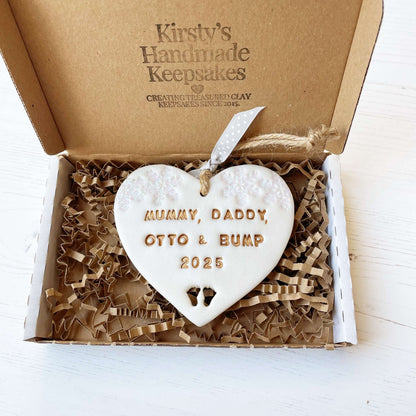 Personalised bumps first Christmas heart ornament, pearlised white clay with MUMMY, DADDY, OTTO & BUMP 2025 painted gold, decorated with 2 iridescent glitter snowflakes on either side of the top of the heart and baby feet cut out at the bottom