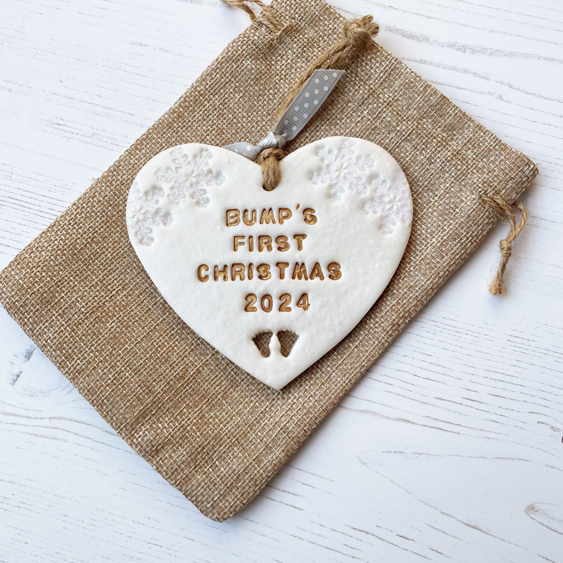 Personalised bump’s first Christmas heart ornament, pearlised white clay with BUMP’S FIRST CHRISTMAS 2024 painted gold, decorated with 2 iridescent glitter snowflakes on either side of the top of the heart and baby feet cut out at the bottom