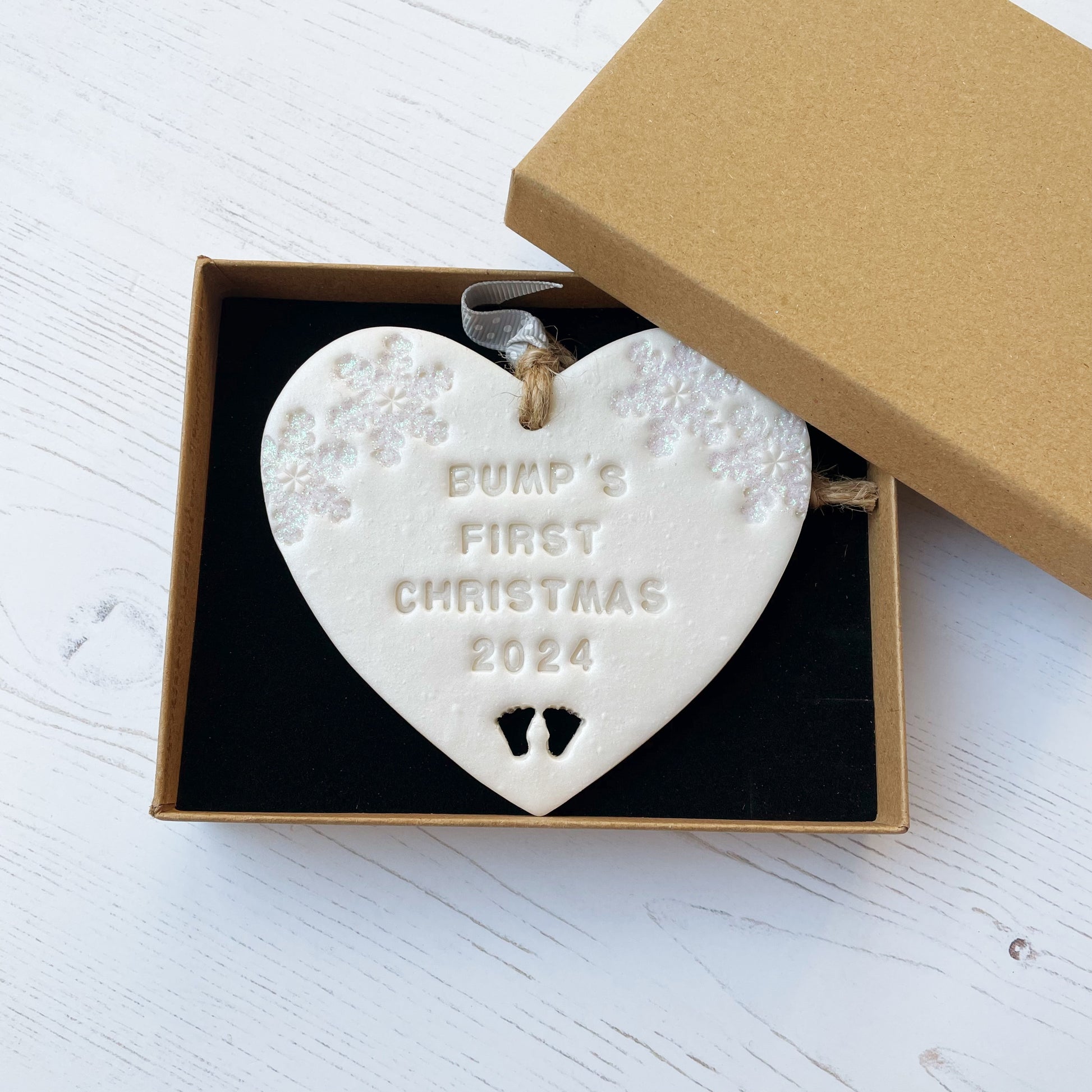 Personalised bump’s first Christmas heart ornament, pearlised white clay with BUMP’S FIRST CHRISTMAS 2024 (not painted), decorated with 2 iridescent glitter snowflakes on either side of the top of the heart and baby feet cut out at the bottom