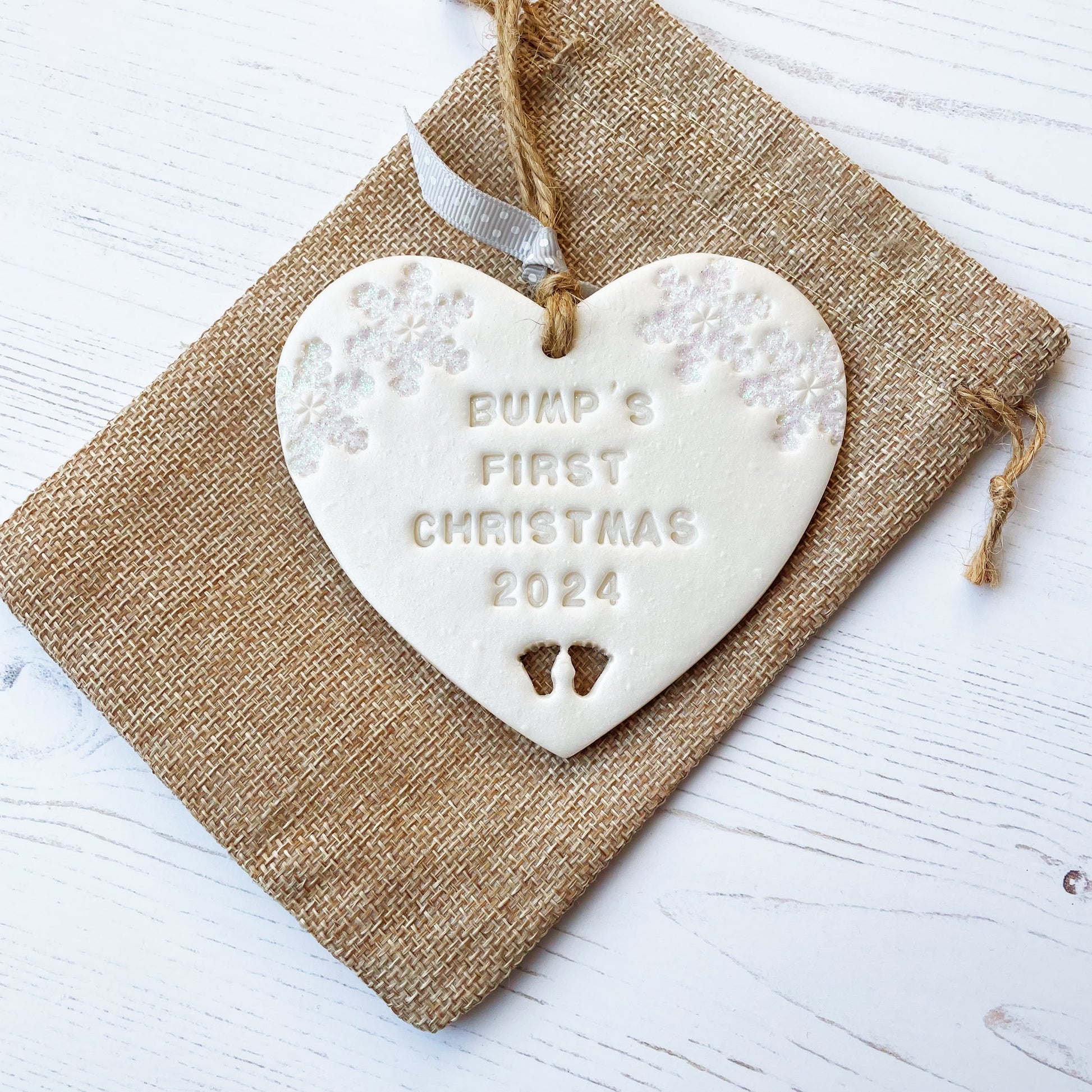 Personalised bump’s first Christmas heart ornament, pearlised white clay with BUMP’S FIRST CHRISTMAS 2024 (not painted), decorated with 2 iridescent glitter snowflakes on either side of the top of the heart and baby feet cut out at the bottom
