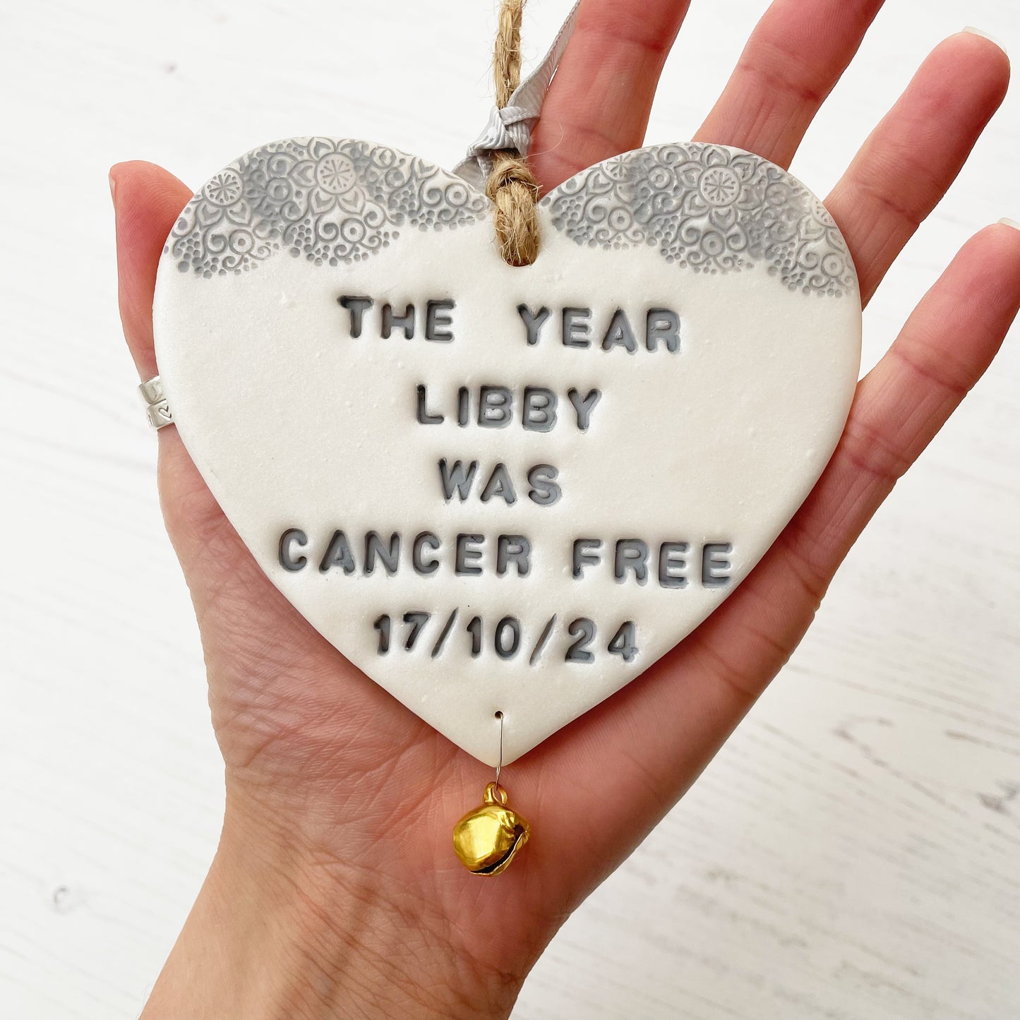 Personalised cancer survivor gift, pearlised white clay hanging heart with a grey lace edge at the top of the heart and a gold bell hanging below, the heart is personalised with THE YEAR LIBBY WAS CANCER FREE 17/10/24