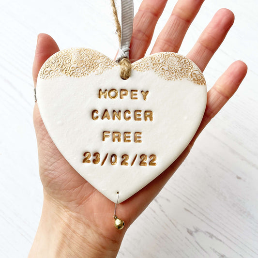 Personalised cancer survivor gift, pearlised white clay hanging heart with a gold lace edge at the top of the heart and a gold bell hanging below, the heart is personalised with HOPEY CANCER FREE 23/02/22